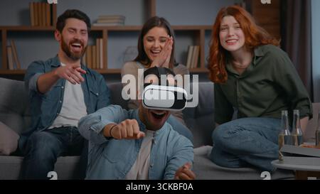 Four multiracial friends at home playing VR game laughing at home party three buddies supporting man in virtual reality helmet play smiling together Stock Photo