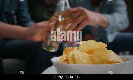 Close up plate with chips snacks four friends clinking bottles on background home party group of multiracial people men women celebrating birthday Stock Photo