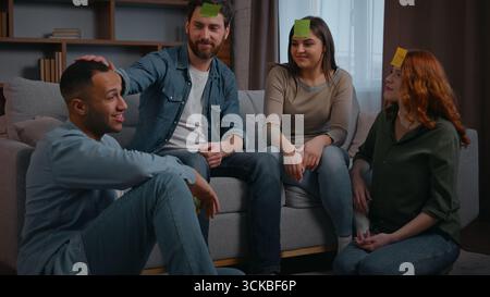 Four diverse friends multiracial people playing guessing game with sticky notes on forehead quess personage word Latina man win buddies applauding Stock Photo