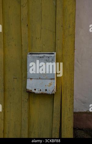 A light blue metal mailbox with rust and wear is mounted on a yellow wooden wall with peeling paint, next to a white plastered section in a rural sett Stock Photo