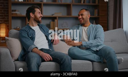 Two multiracial men multiethnic guys talking friendly at home relaxing on couch sofa communicate Caucasian American male friendship discuss funny news Stock Photo