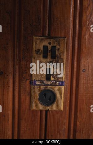 A detailed view of an old electrical switch panel with black switches and a round socket, mounted on a grooved wooden wall in a rustic setting. Stock Photo
