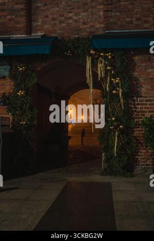 A nighttime view in Gdansk showing a brick archway with festive greenery and ornaments, a dimly lit cobblestone path, and atmospheric fog. Stock Photo