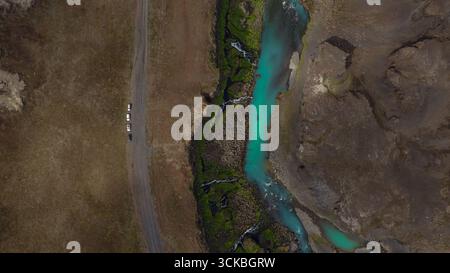 Turquoise river flows through rugged Icelandic landscape with green moss, small waterfalls, barren terrain, and a narrow road with parked vehicles. Stock Photo