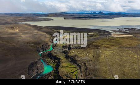 A turquoise river flows through a deep canyon in Iceland, surrounded by earthy terrain, a large lake, a winding road, and distant mountains. Stock Photo