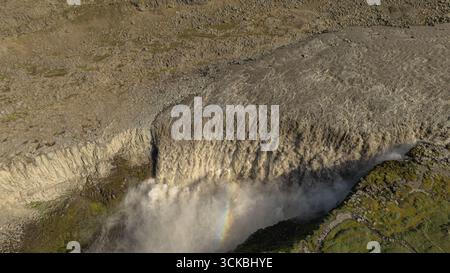 Dramatic aerial view of Dettifoss waterfall cascading over a rugged cliff with mist, a faint rainbow, and rocky terrain in Vatnajokull National Park. Stock Photo