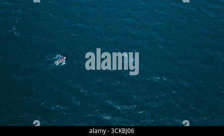 Aerial perspective of a small boat with passengers whale watching in deep blue ocean waters, with a whale partially visible beneath the surface. Stock Photo