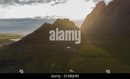 Dramatic aerial view of Icelandic mountains with sunlight streaming through peaks, a small building, green fields, a winding road, and distant water. Stock Photo