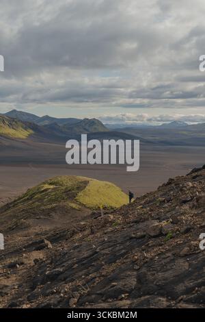 A lone figure stands near a grassy hill in a rugged volcanic terrain. Dark rocks, vibrant green moss, and distant mountains create a dramatic scene. Stock Photo