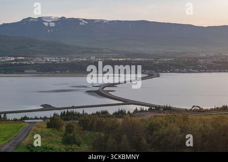 Akureyri in northern Iceland features a curved causeway, snow capped mountains, lush greenery, and a fjord reflecting soft sunlight. A construction ve Stock Photo