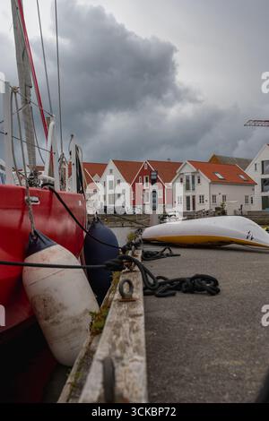 A red sailboat with visible fenders and mooring lines rests at a dock. Nearby are white and red roofed houses, with gray clouds overhead in a Norwegia Stock Photo