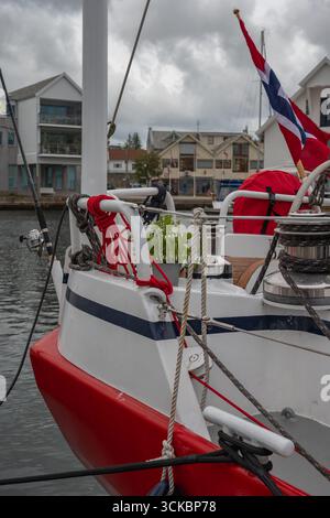 A red and white sailboat with coiled ropes and a Norwegian flag docked in a calm harbor. Waterfront buildings and an overcast sky form the backdrop. Stock Photo
