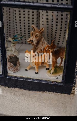 A black framed window showcases ceramic figurines of a cow, deer, wolf, bird, and starfish on a white surface with dotted white curtains behind. Stock Photo
