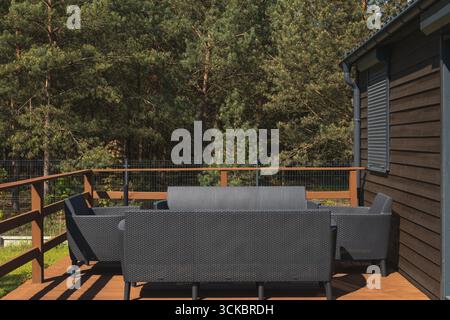 A wooden deck attached to a dark brown cottage, featuring black woven furniture and a metal mesh railing, surrounded by tall pine trees in sunlight. Stock Photo