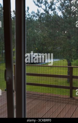Scene from inside a house showing a wooden deck, a black Tesla car on a driveway, grassy yard, tall pine trees, and a metal fence in the distance. Stock Photo