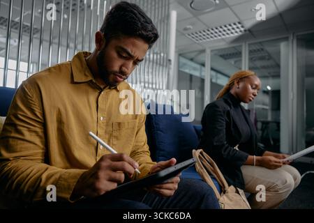 Two multiracial young adults in businesswear sitting on sofa and writing on digital tablet in office Stock Photo