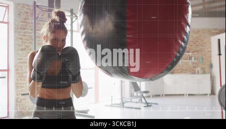 Woman boxer wearing sports bra throwing punches at punching bag in gym studio, with boxing gloves. Fitness, strength, determination, athleticism, trai Stock Photo