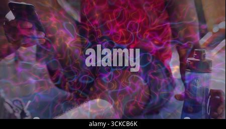 Sitting man wearing red top holding smartphone and blue vape device on couch, neon light trails. Neon, digital, technology, lounge, vibrant, relaxatio Stock Photo