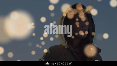 Standing adult female wearing hooded parka outdoors against dusk sky, with warm-toned bokeh overlay. Nature, serenity, tranquility, dusk, outdoor, fas Stock Photo