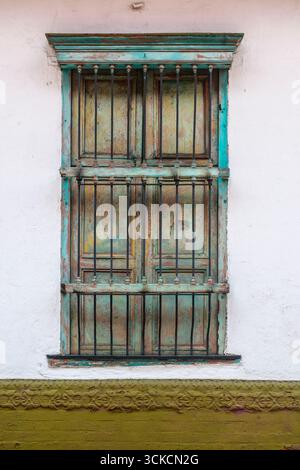 Weathered, mainly turquoise colored, window with iron bars and closed shutters in a whitewashed wall. Cali, Colombia Stock Photo
