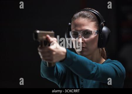 Caucasian young adult woman wearing protective glasses and earmuffs aiming handgun at shooting range, focusing intently on target with both hands extended forward Stock Photo