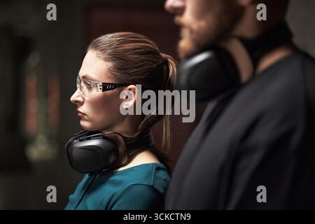 Caucasian young woman wearing protective glasses and earmuffs standing next to Caucasian young man with beard also wearing earmuffs, both focusing on task in industrial setting Stock Photo