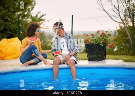 Silly couple sitting by the pool in swimming goggles eating watermelon fruit and talking. Lifestyle, outdoors, togetherness concept. Stock Photo