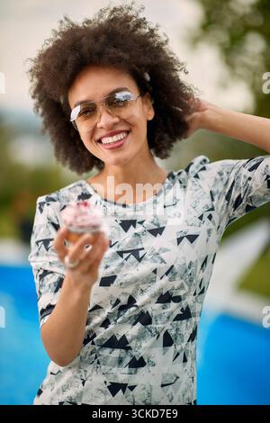 Happy young woman feeling joyful and posing outdoors, holding a cupcake. Lifestyle, fun, joy concept. Stock Photo