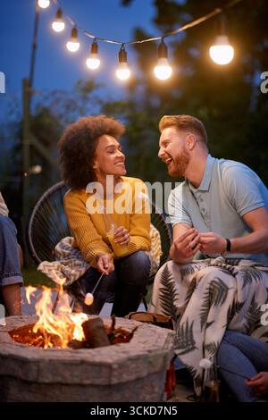 Attractive young couple sharing a romantic moment together, sitting by the fire roasting marshmallows on a summer evening. Togetherness, joy, fun conc Stock Photo