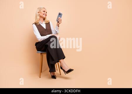 Beautiful senior businesswoman in formal attire using smartphone while sitting against plain beige background Stock Photo