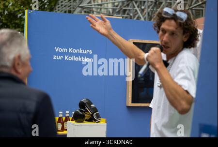 Munich, Germany. 11th Sep, 2025. Performance artist Jakob Margit Wirth (r) talks to a passer-by during an art campaign in the city center. In the background you can see boxing gloves and ketchup bottles standing and lying in front of a wall with the inscription "From bankruptcy to collection". Action artists have opened a "pop-up store" in front of the Alte Akademie, where the possessions of the failed Austrian real estate tycoon Benko are to be sold. Credit: Leonie Asendorpf/dpa/Ģ����ý Live News Stock Photo