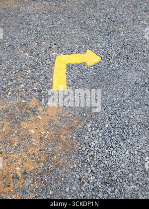 Close-up view of a faded yellow arrow on a gravel-covered parking area, pointing right with a bend, on rough stones and dirt. Perfect for illustrating Stock Photo