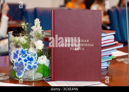 Russian graduation thesis book on academic table with floral arrangement and stacked documents. Stock Photo
