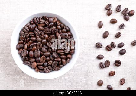 Roasted  Arabica coffee beans, in a white bowl, on linen. Dark brown roasted whole seeds of berries from Coffea arabica. Stock Photo