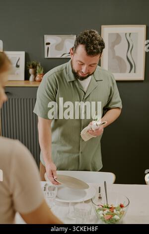 Caucasian Young Adult Man Setting Table and Polishing Plate before Meal Stock Photo