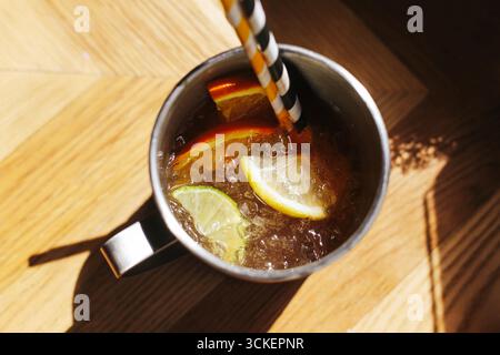 Refreshing cold drink with lemon slices. Summer beverage in metal mug. Ice cubes with citrus on wooden table. Stock Photo