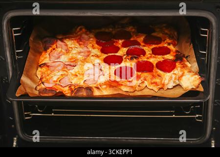Homemade pizza with salami and cheese. Fresh pizza baking in oven. Delicious Italian food ready to eat. Crispy golden crust with toppings. Stock Photo