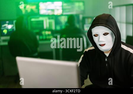 Hacker working on laptop in dark room with green code displayed on monitors Stock Photo