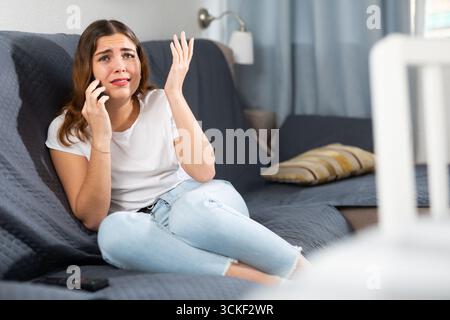 Dissatisfied young woman complaining to anyone on mobile phone while sitting on sofa Stock Photo