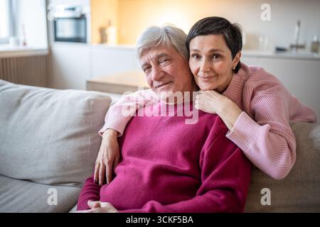 Smiling senior married couple hugging on sofa at home looking at camera. Happy family love and care Stock Photo
