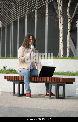 Confident business man sitting on bench with laptop in park. Young businessman enjoying work break. Businessman taking break on park bench. Stock Photo