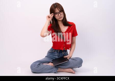 A beautiful young woman wearing glasses and casual attire sits cross-legged while reading in studio background Stock Photo