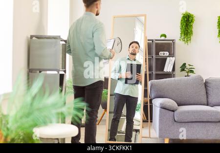 Young man looking at the mirror preparing for work standing at home and holding documents in hands Stock Photo