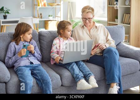Happy family living room, senior woman and two kids on sofa, using phone, looking at laptop screen Stock Photo