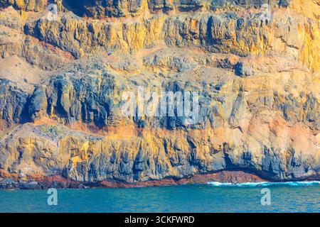 Towering rock formation rises dramatically from calm ocean. Rugged cliff face shows distinct layers and textures, with water visible below Stock Photo