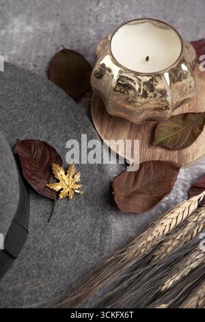 Moody autumn still life: grey felt hat with gold leaf brooch, pumpkin‑shaped candle on a wooden board, burgundy leaves, and wheat stalks. Stock Photo