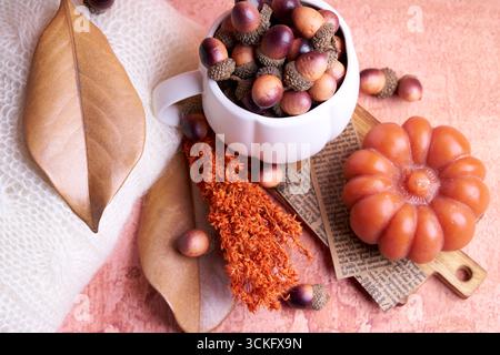 Cozy autumn still life: pumpkin‑shaped white mug of acorns, pumpkin candle on a cutting board, dried leaves and orange sprig on a peach background. Stock Photo