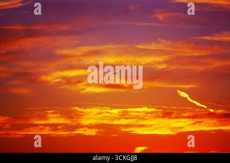 Dramatic sky at sunset with airplane silhouette flying through colorful clouds. Fiery tones of orange, yellow and red blend creating breathtaking view Stock Photo