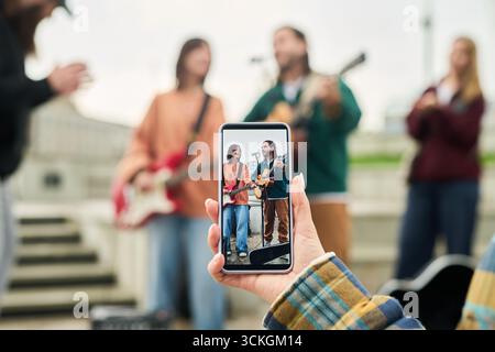 Caucasian young adult woman holding smartphone capturing group of young adult musicians performing outdoors, multiethnic band members playing instruments, focus on phone screen Stock Photo
