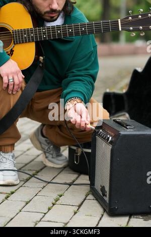 Young adult Caucasian man kneeling outdoors connecting electric guitar to amplifier, holding instrument with one hand and plugging cable with other, preparing for performance Stock Photo